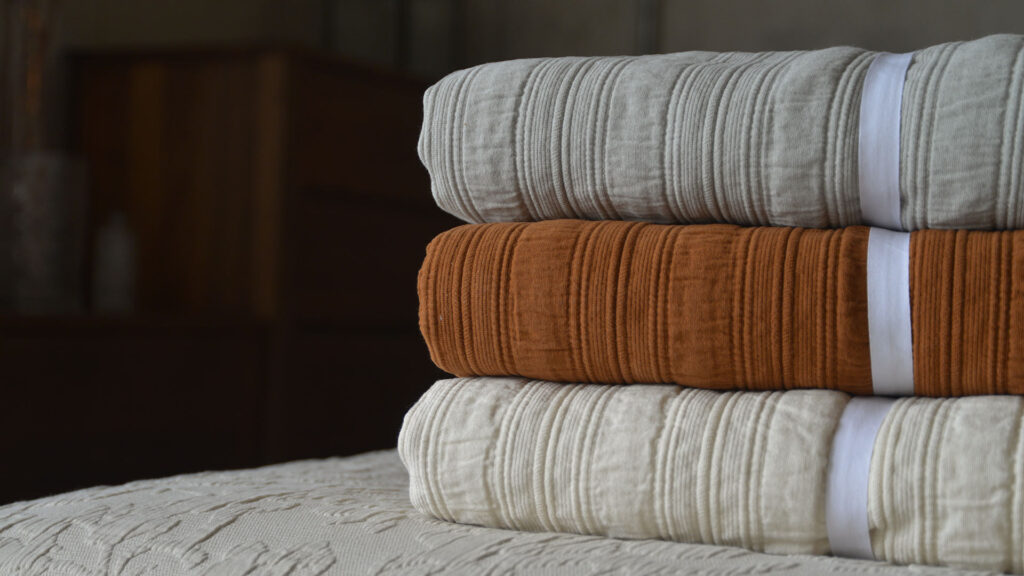 Stack of folded stripe cotton bedspreads, in the colours silver grey (top), cinnamon (middle) and ivory (bottom).