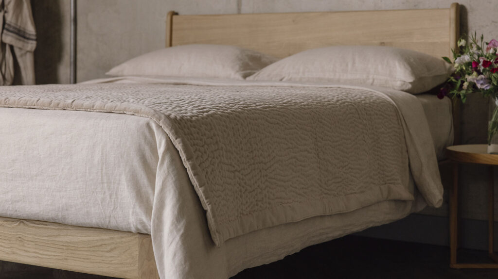 Close up shot of the the stitched velvet bedspread in soft beige and the organic ecru linen bedding on top of the English oak Camden.