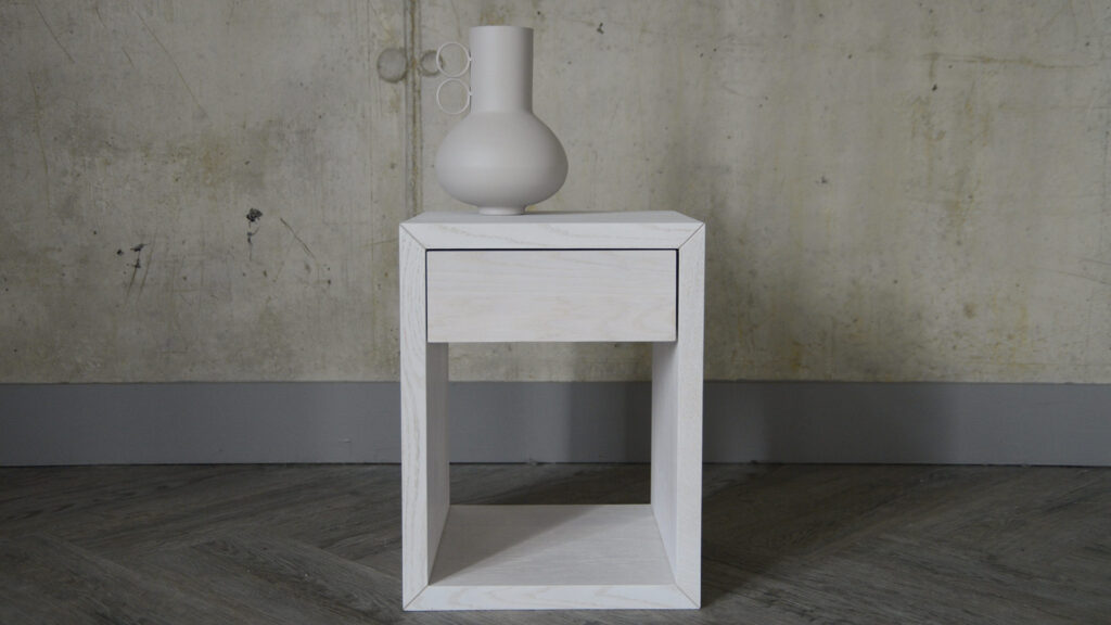 Image showing a small bedside table stained with a pure white finish. A decorative vase in a similar pale colour has been placed atop it.