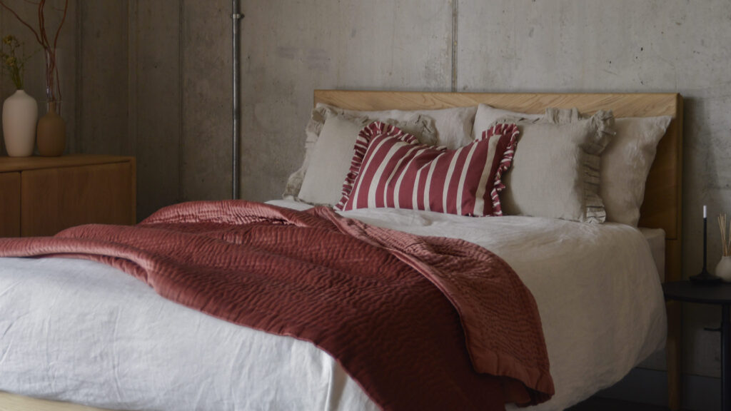 Oak bed frame with ecru bed linen, a wine striped cushion and a wine coloured velvet bedspread.